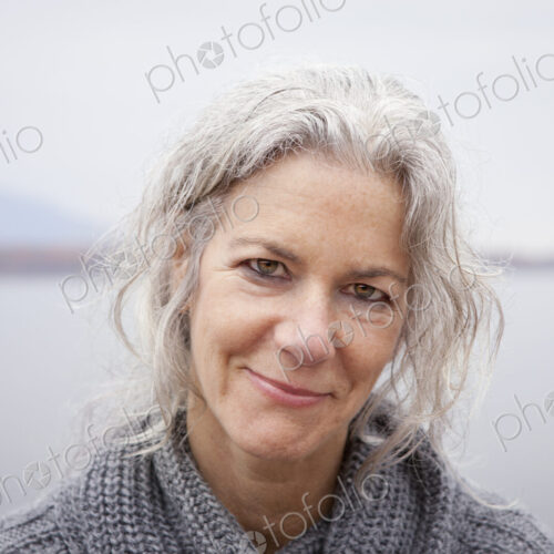 A woman looking at the camera, by the shore of a lake.