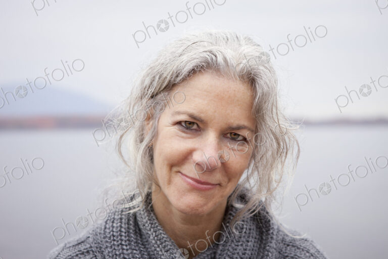 A woman looking at the camera, by the shore of a lake.