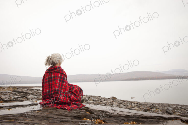 A woman looking out over the water on the shores of a calm lake, wrapped in a tartan rug.
