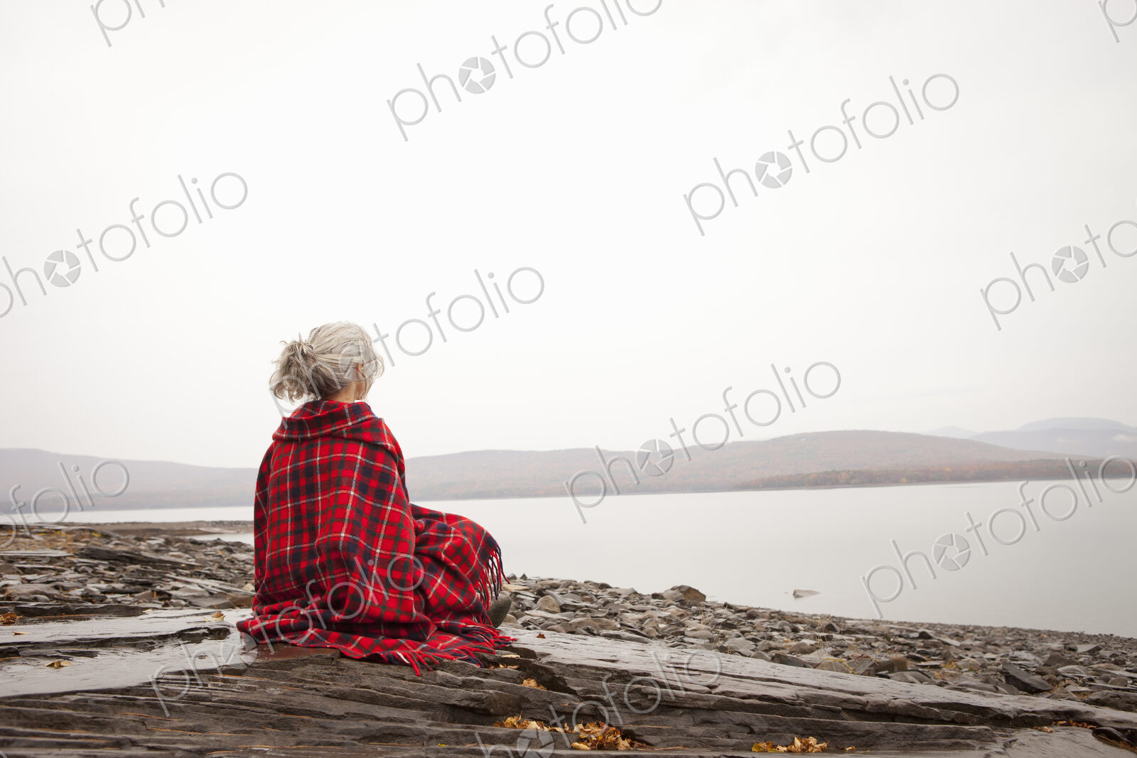 A woman looking out over the water on the shores of a calm lake, wrapped in a tartan rug.
