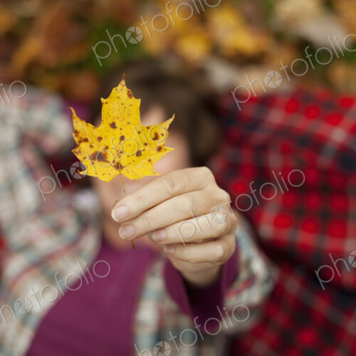 A woman lying on a red tartan picnic blanket, looking upwards, holding a maple leaf. A woman lying on a red tartan picnic blanket, looking upwards, holding a maple leaf.