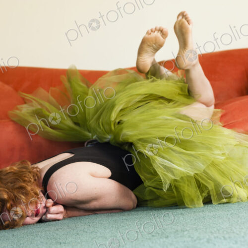 A woman lying on her front on the floor, wearing a lime green tutu, kicking her legs…