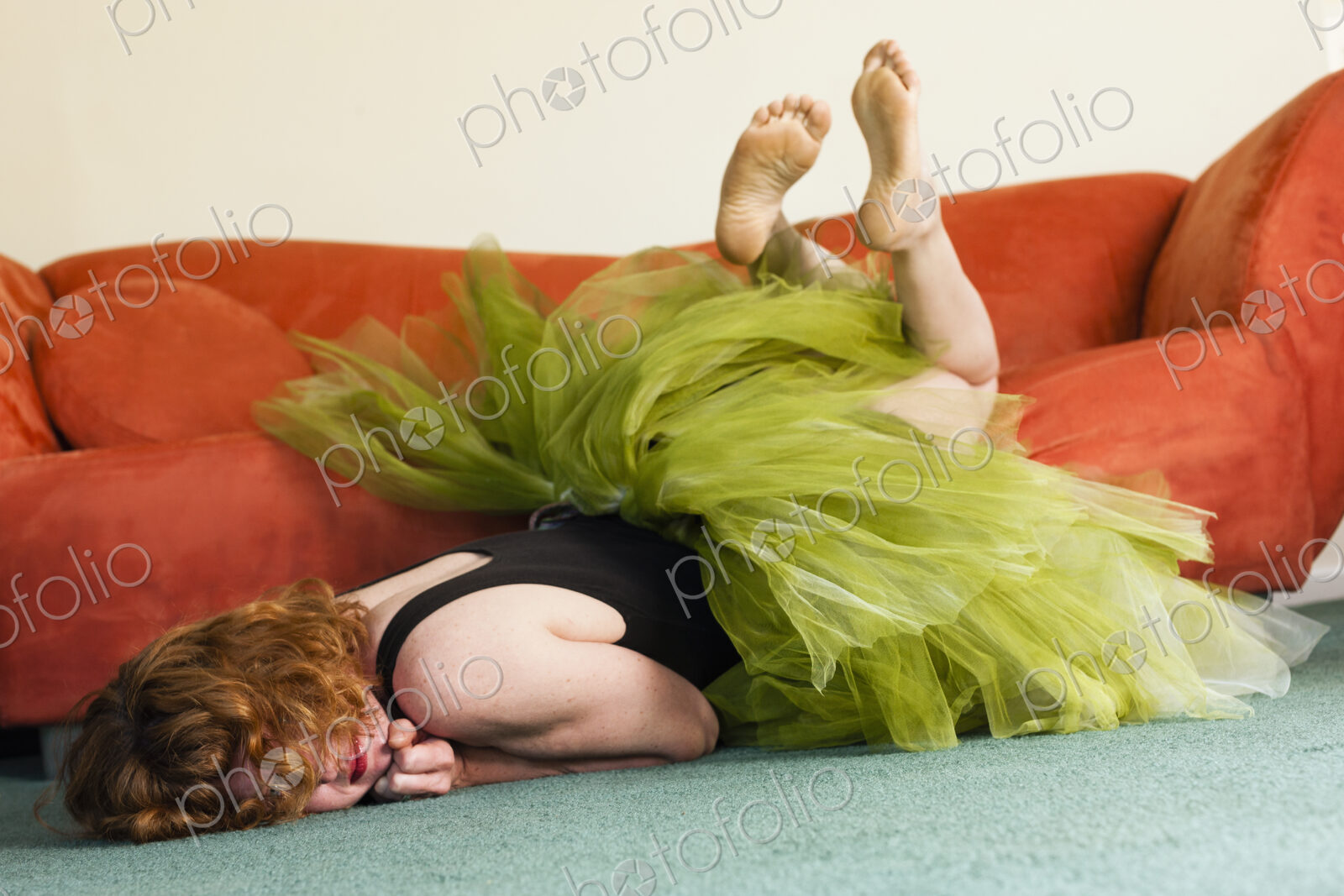 A woman lying on her front on the floor, wearing a lime green tutu, kicking her legs…