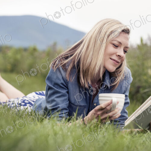 A woman lying on the grass holding a tea cup and reading a book. A woman lying on the grass holding a tea cup and reading a book.