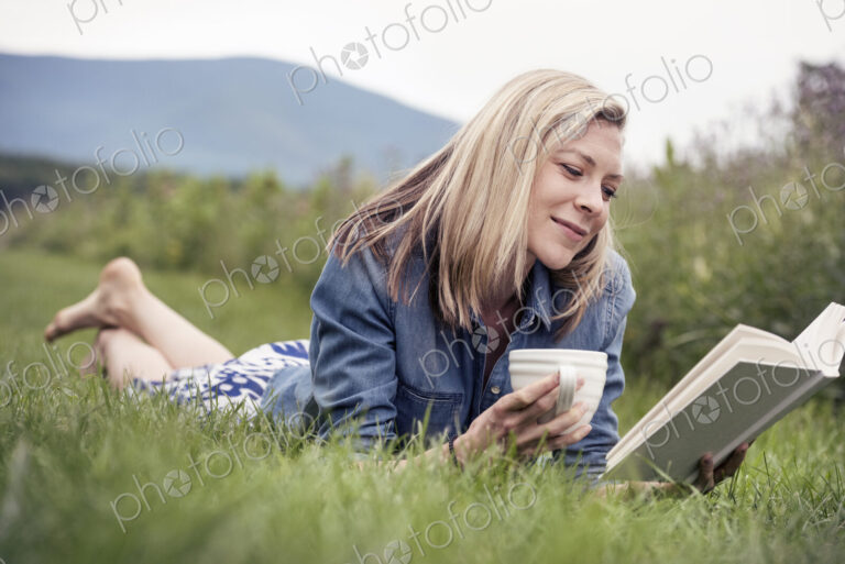 A woman lying on the grass holding a tea cup and reading a book.