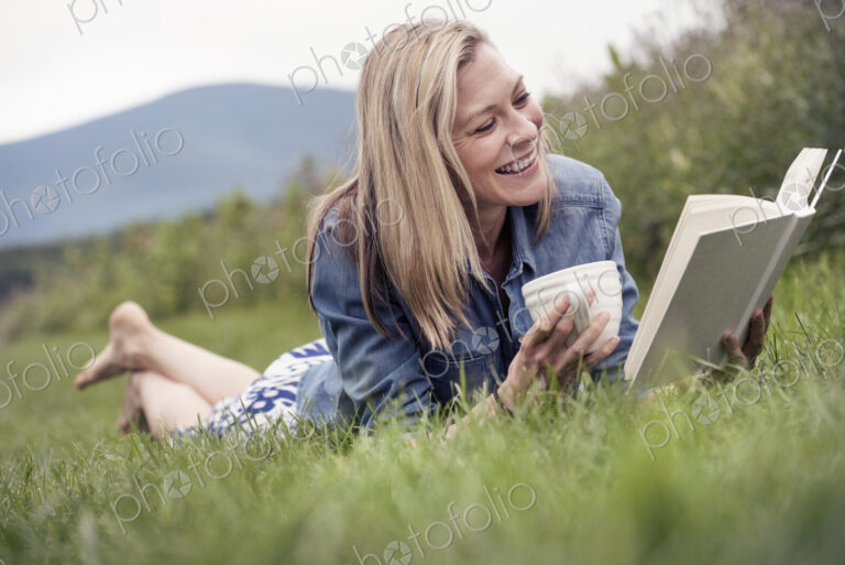 A woman lying on the grass holding a tea cup and reading a book.