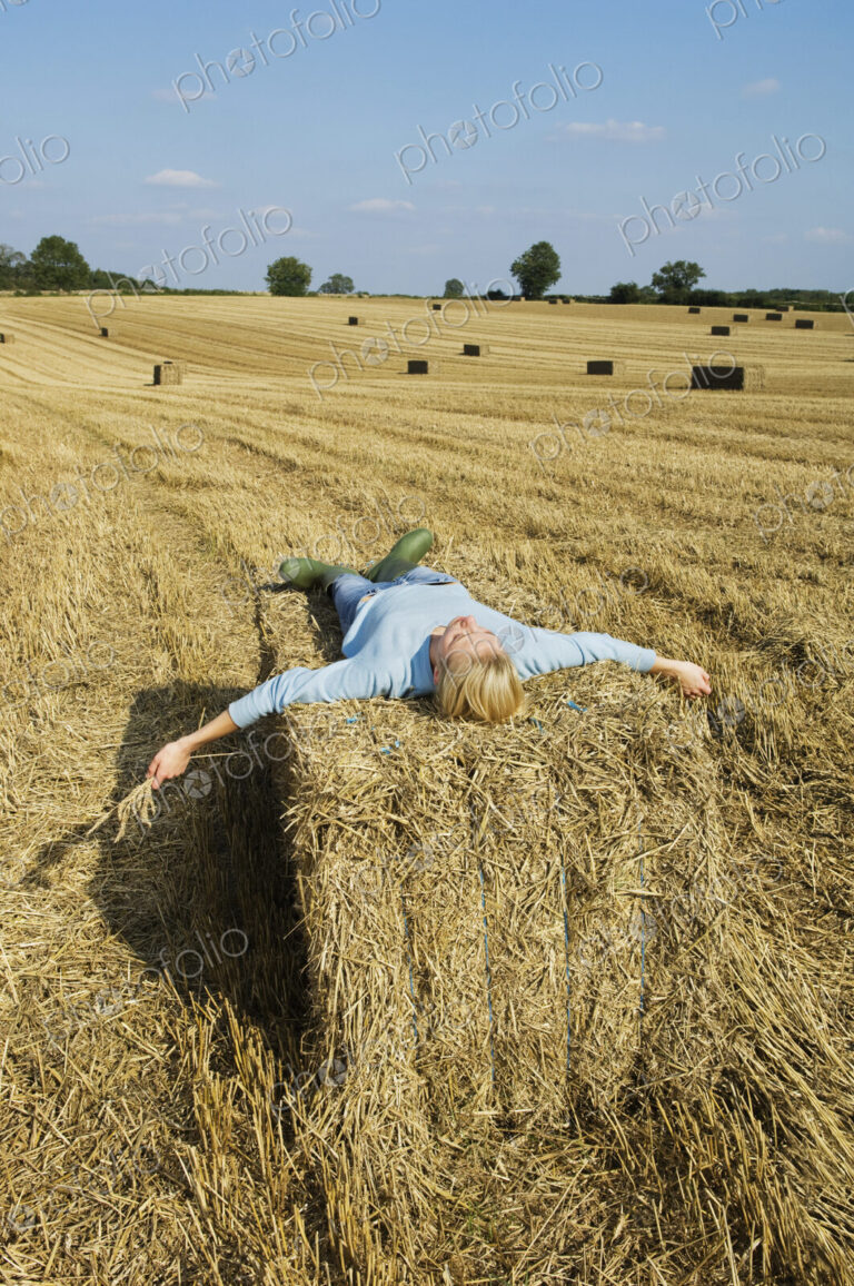 A woman lying on top of a square bale of packed straw at harvest time.