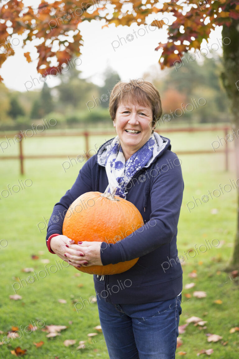 A woman outdoors holding a large orange pumpkin.