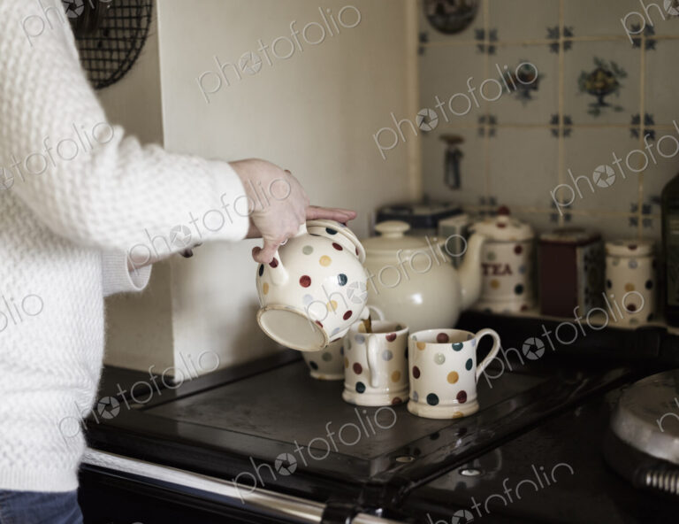 A woman pouring tea, by a range cooker. Using decorative china.