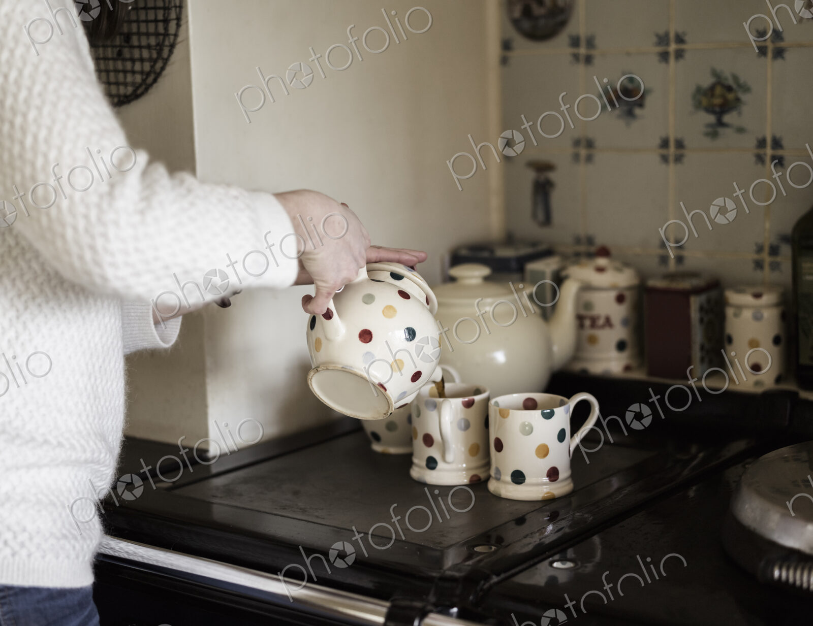 A woman pouring tea, by a range cooker. Using decorative china.