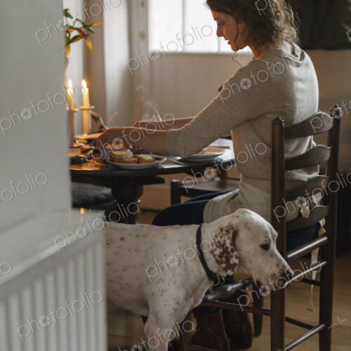 A woman seated at a table in a ladderback chiar, a large white dog beside her. A woman seated at a table in a ladderback chiar, a large white dog beside her.