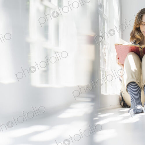 A woman seated by a window, reading with a book on her lap. A woman seated by a window, reading with a book on her lap.