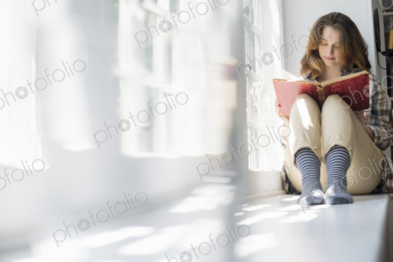 A woman seated by a window, reading with a book on her lap.