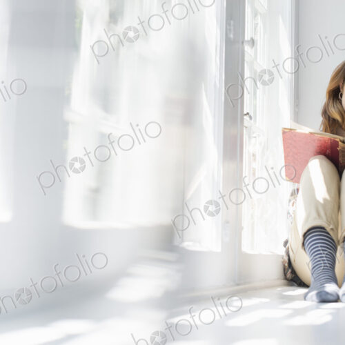 A woman seated by a window, reading with a book on her lap. A woman seated by a window, reading with a book on her lap.