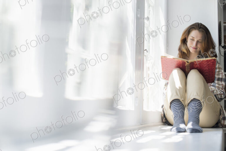 A woman seated by a window, reading with a book on her lap.
