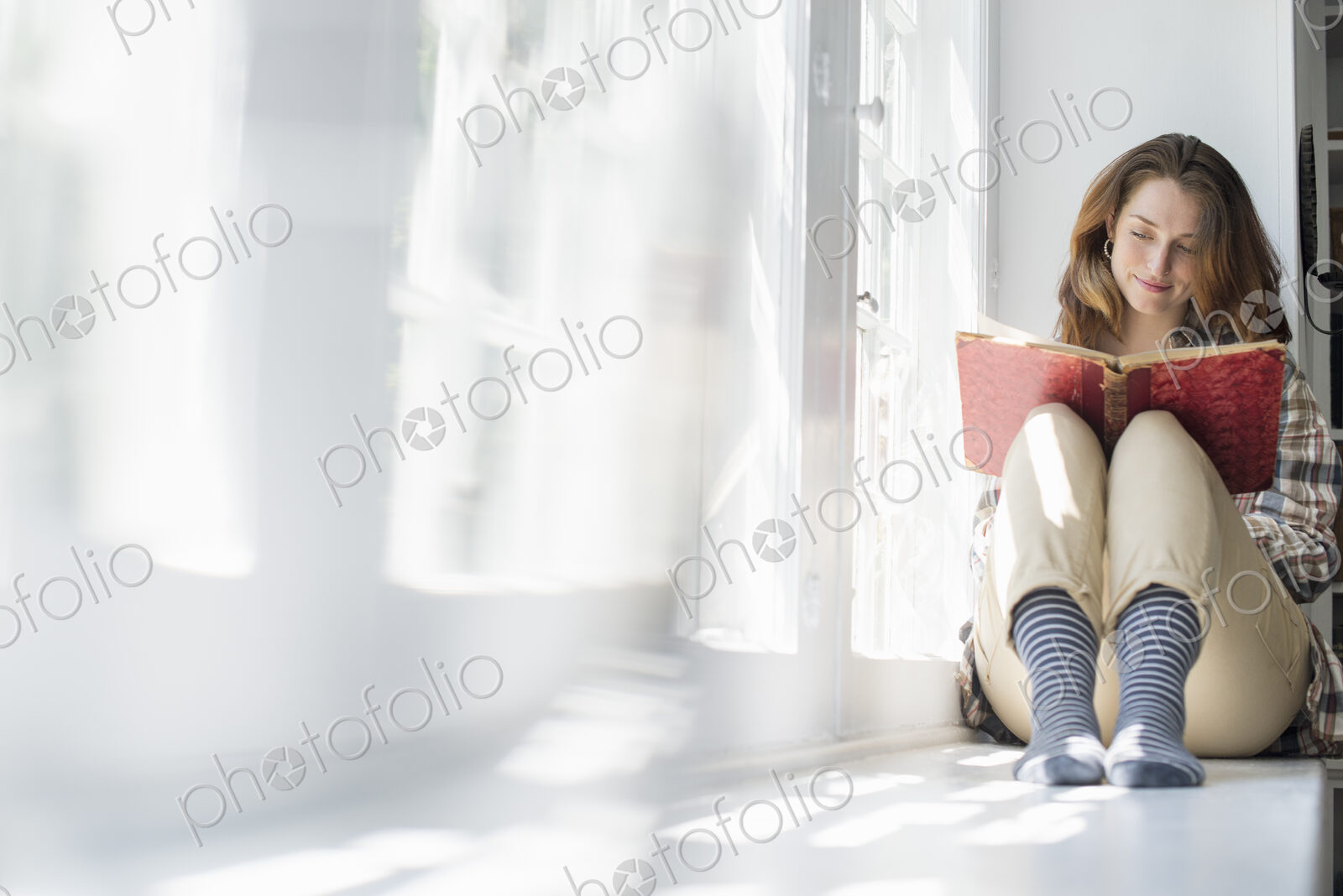 A woman seated by a window, reading with a book on her lap.