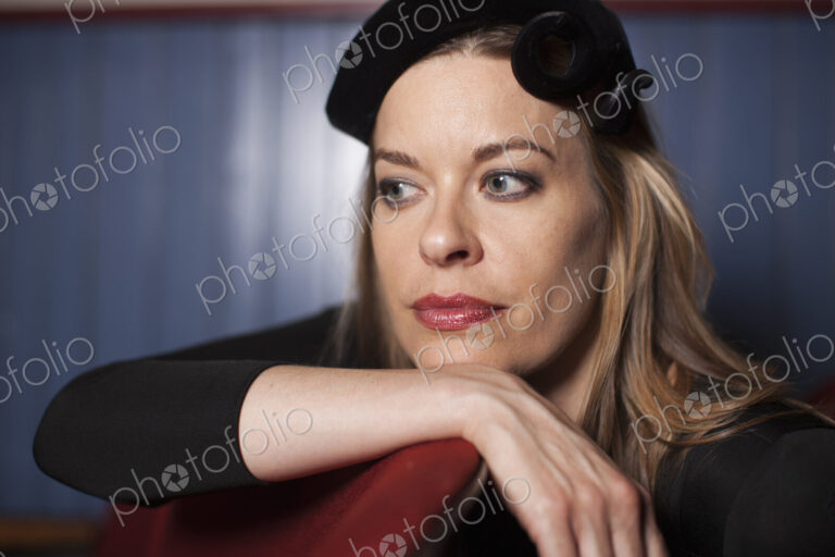 A woman seated in a theatre, wearing an elegant evening hat, and a black top. Resting…