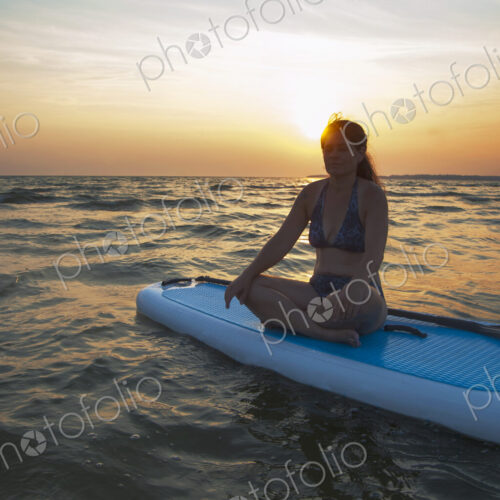 A woman seated on a paddleboard looking out at the sun setting on the horizon. A woman seated on a paddleboard looking out at the sun setting on the horizon.