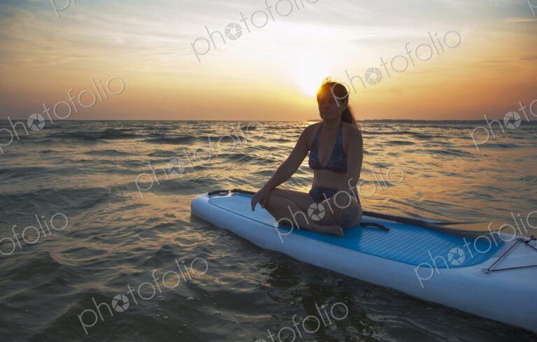 A woman seated on a paddleboard looking out at the sun setting on the horizon.