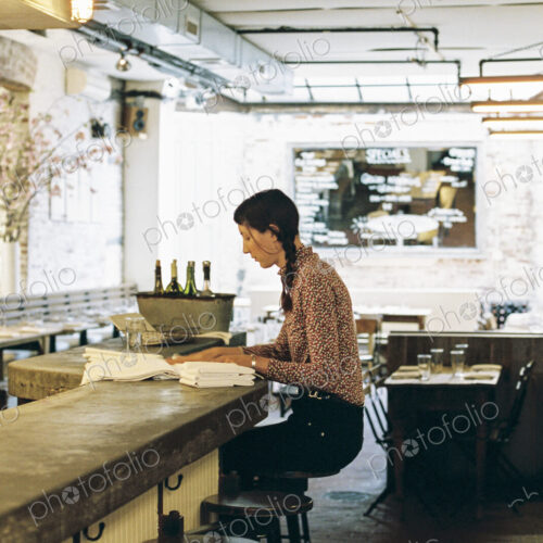 A woman seated on a tall stool, folding napkins for a restaurant table. A woman seated on a tall stool, folding napkins for a restaurant table.