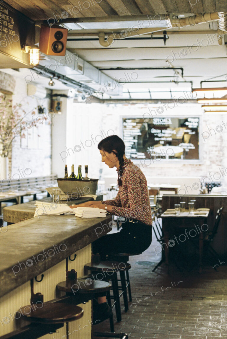 A woman seated on a tall stool, folding napkins for a restaurant table.
