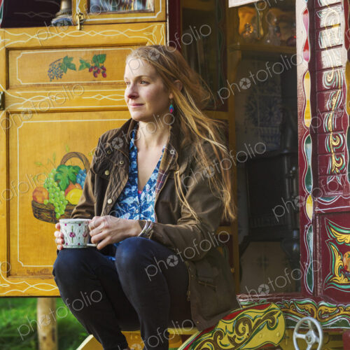 A woman seated on the steps of a caravan, holding a mug of tea.