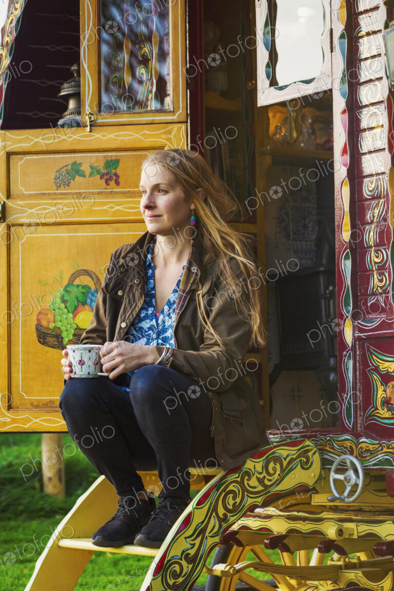 A woman seated on the steps of a caravan, holding a mug of tea.