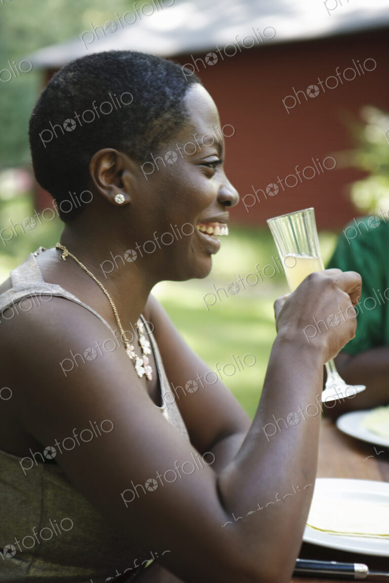 A woman sitting at a table outdoors in a garden, taking a drink.
