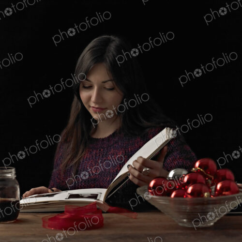A woman sitting at a table reading a book. A glass bowl of red shiny baubles, and a… A woman sitting at a table reading a book. A glass bowl of red shiny baubles, and a…