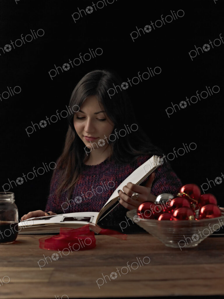 A woman sitting at a table reading a book. A glass bowl of red shiny baubles, and a…