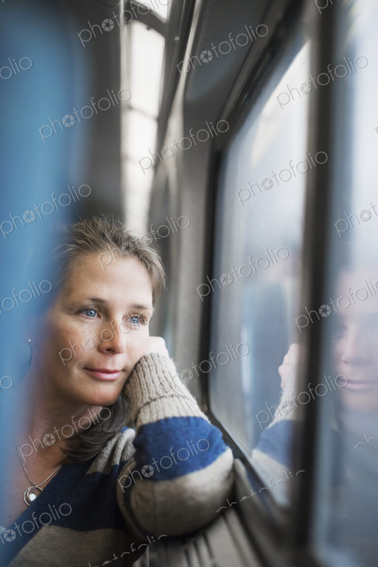 A woman sitting at a window seat in a train carriage, resting her head on her hand.…