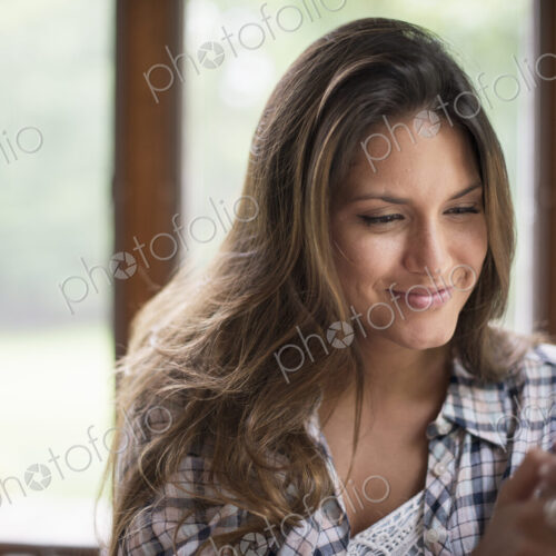 A woman sitting by a window, looking at her cell phone.