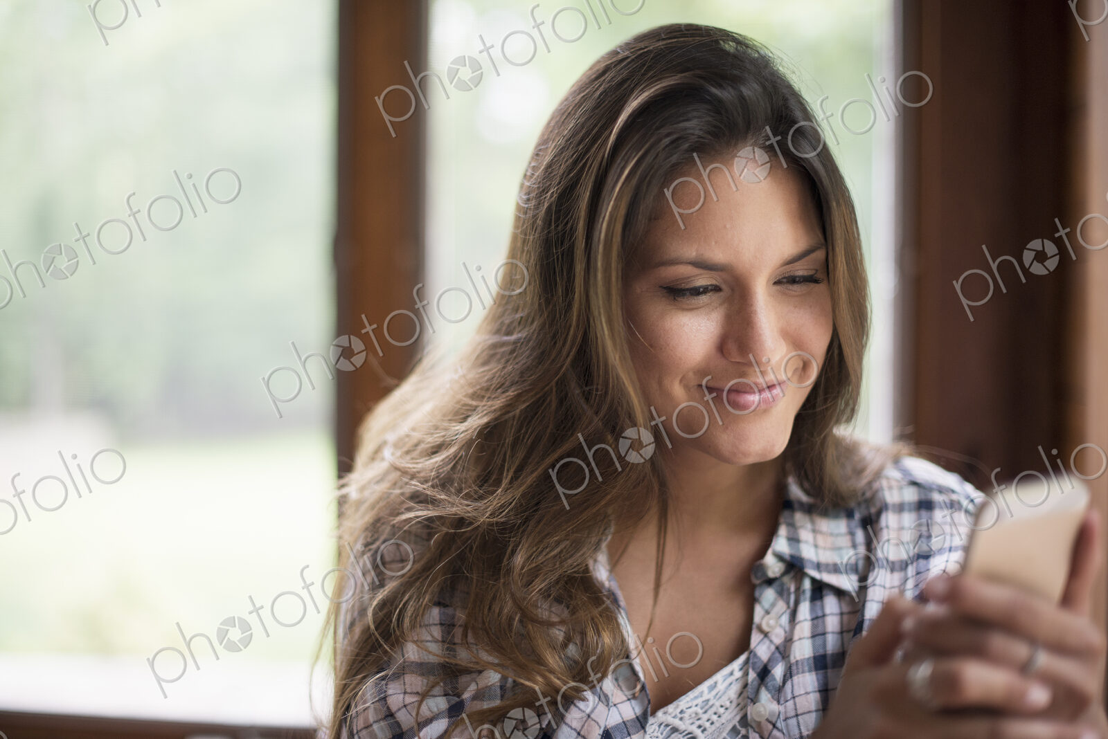 A woman sitting by a window, looking at her cell phone.
