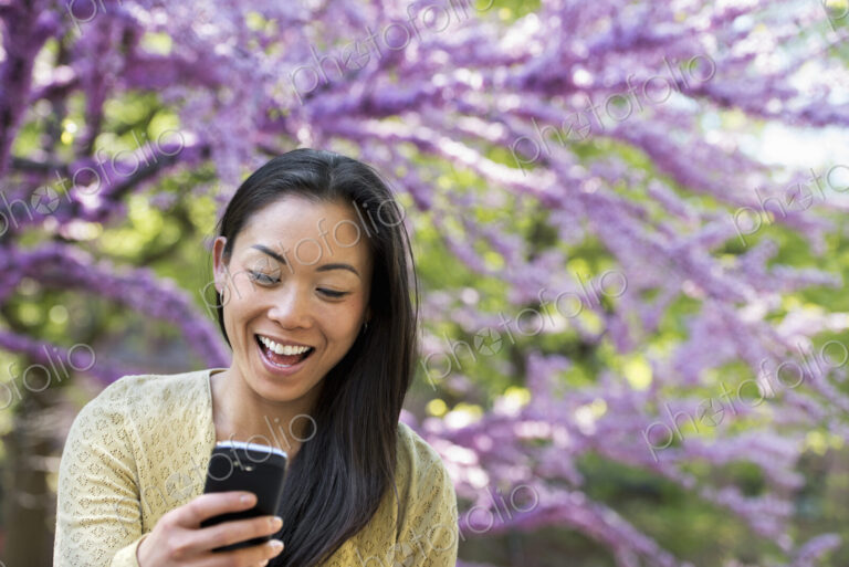 A woman sitting in a park under a tree with pink blossom. Looking at her smart phone…
