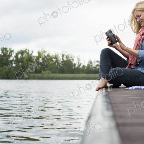 A woman sitting on a jetty by a lake, using a digital tablet. A woman sitting on a jetty by a lake, using a digital tablet.