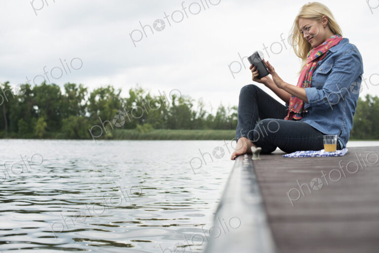 A woman sitting on a jetty by a lake, using a digital tablet.