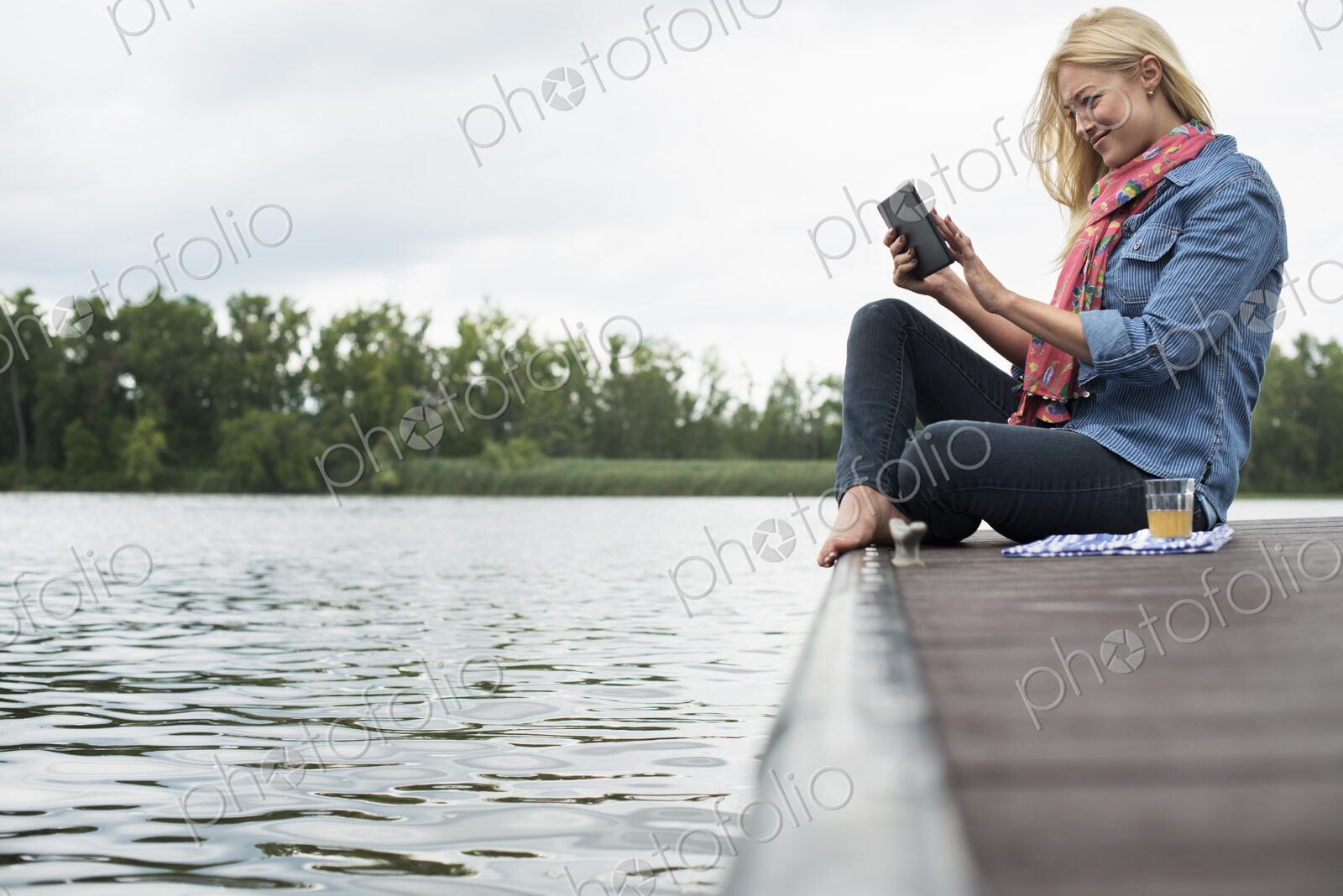 A woman sitting on a jetty by a lake, using a digital tablet.