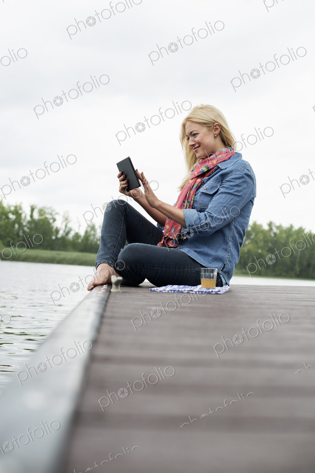 A woman sitting on a jetty by a lake, using a digital tablet.