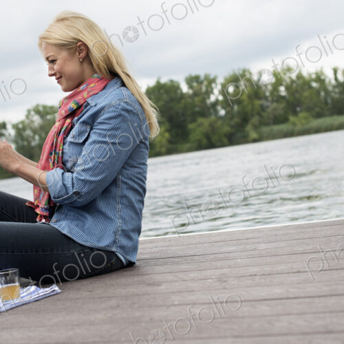 A woman sitting on a jetty by a lake, using a digital tablet.