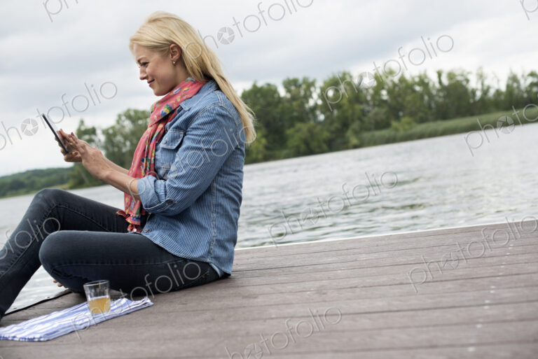 A woman sitting on a jetty by a lake, using a digital tablet.