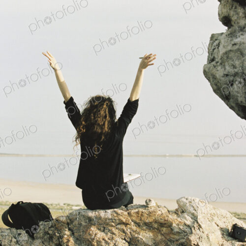 A woman sitting on a rock with her arms raised, looking out over water