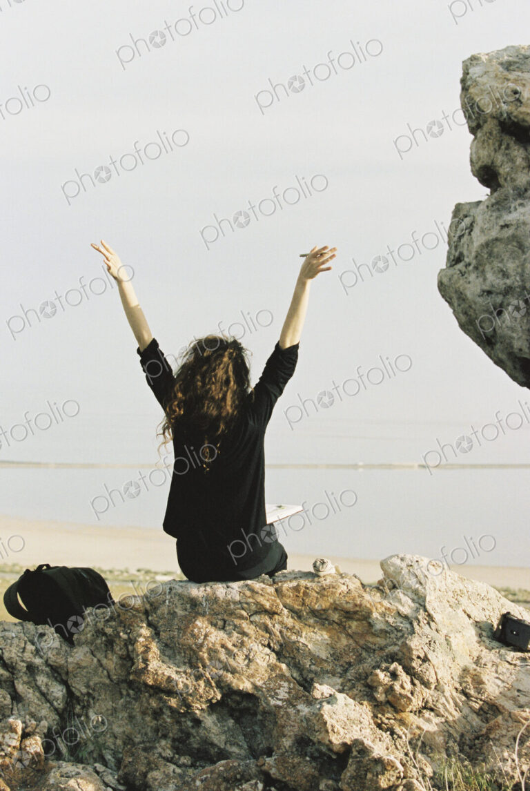 A woman sitting on a rock with her arms raised, looking out over water