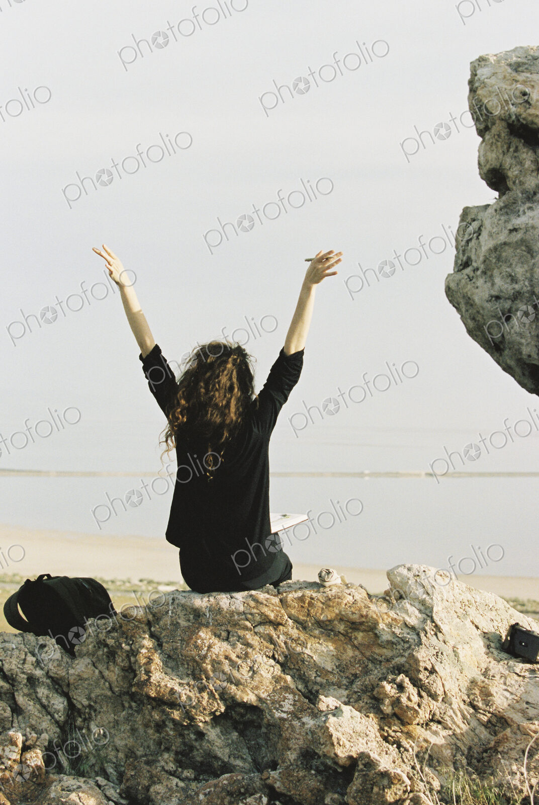 A woman sitting on a rock with her arms raised, looking out over water