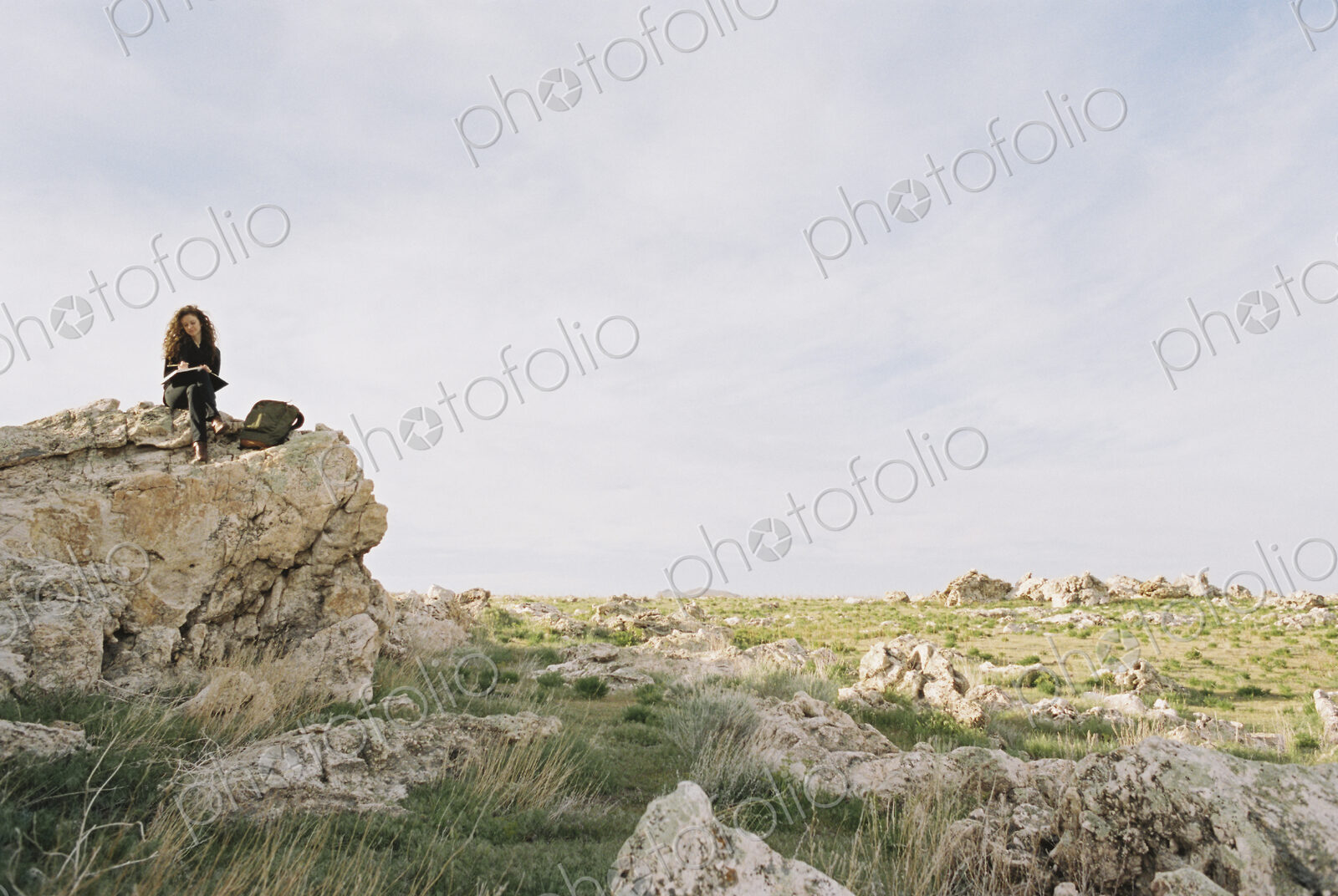 A woman sitting on a rock, writing in a notebook, a backpack lying by her feet.