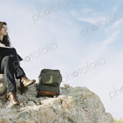 A woman sitting on a rock, writing in a notebook, a backpack lying by her feet. A woman sitting on a rock, writing in a notebook, a backpack lying by her feet.