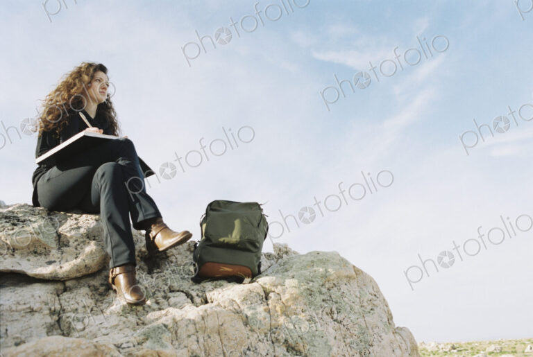 A woman sitting on a rock, writing in a notebook, a backpack lying by her feet.