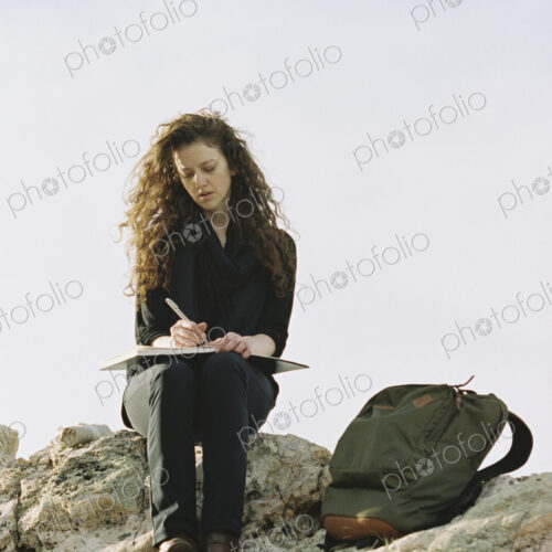 A woman sitting on a rock, writing in a notebook, a backpack lying by her feet.