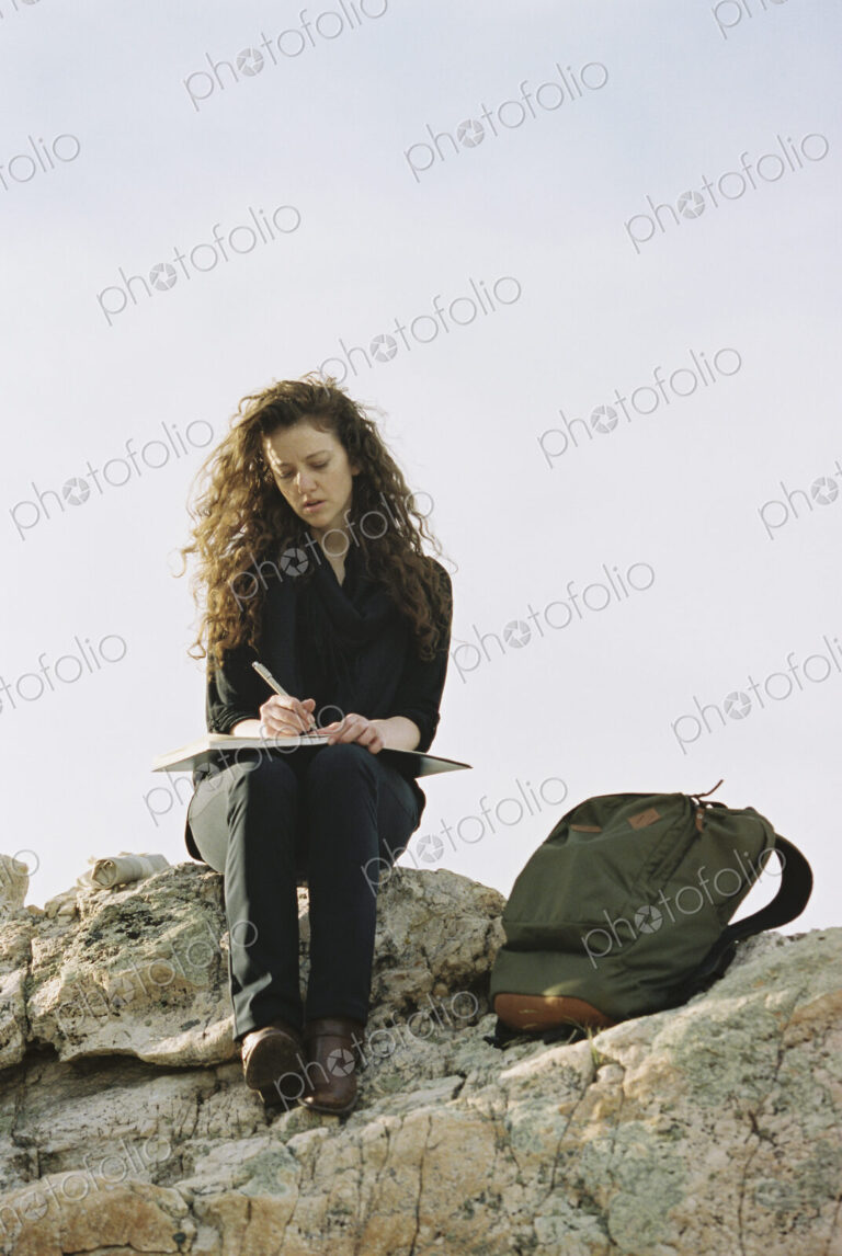 A woman sitting on a rock, writing in a notebook, a backpack lying by her feet.