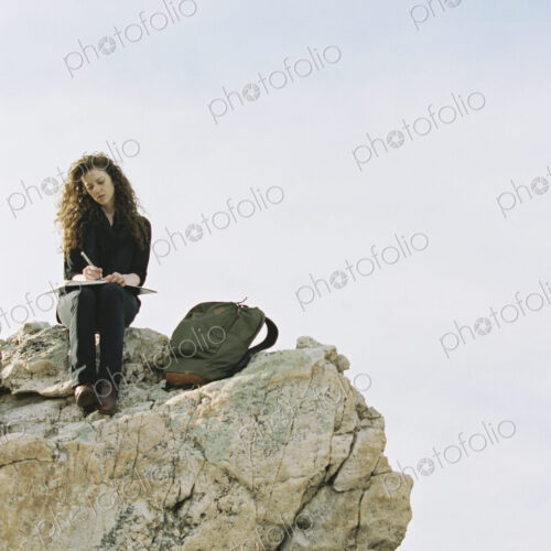 A woman sitting on a rock, writing in a notebook, a backpack lying by her feet.