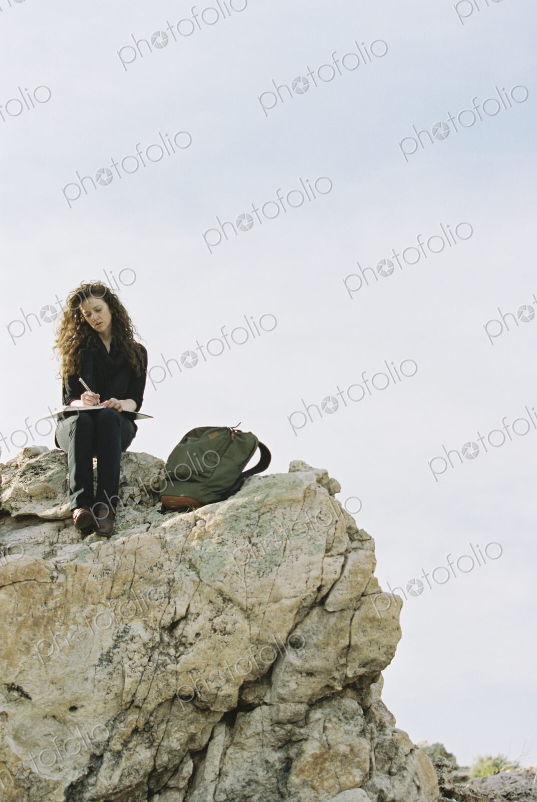 A woman sitting on a rock, writing in a notebook, a backpack lying by her feet.